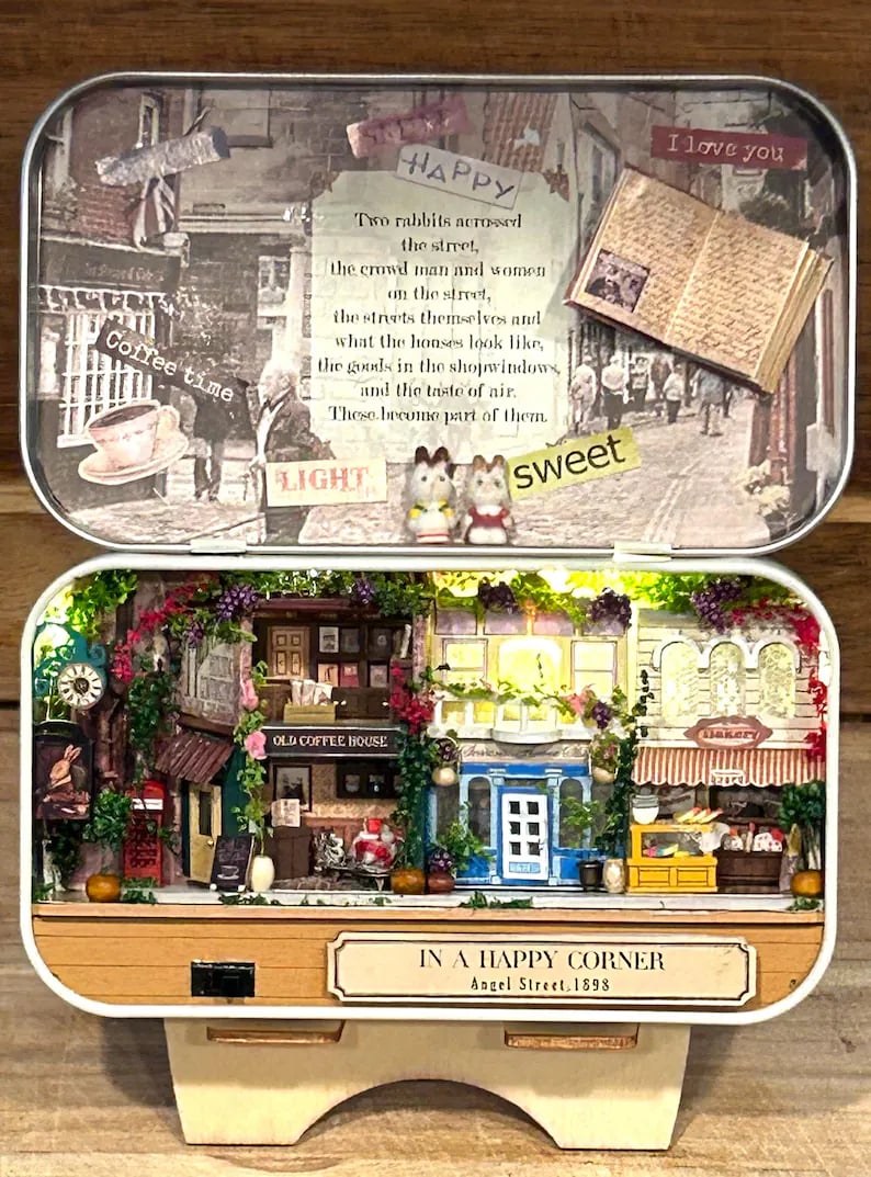 可能包括：一个街景微缩模型，其中有两家商店，一家标有“老咖啡馆”，另一家标有“面包店”。模型由木头制成，有一个白色底座和一个木架。模型从内部照明，中央有一个小灯泡。模型周围绿树环绕，左侧有一个小钟。模型放在一个带盖的锡盒里，盒子里有一幅街景图，上面写着“快乐”和“我爱你”。盖子上的文字写着：“街对面的两只兔子，街上拥挤的男人和女人，街道本身和房屋的样子，商店橱窗里的商品，以及空气的味道。这些都成为它们的一部分。”模型底座上的文字写着：“在快乐的角落，天使街，1898 年。”